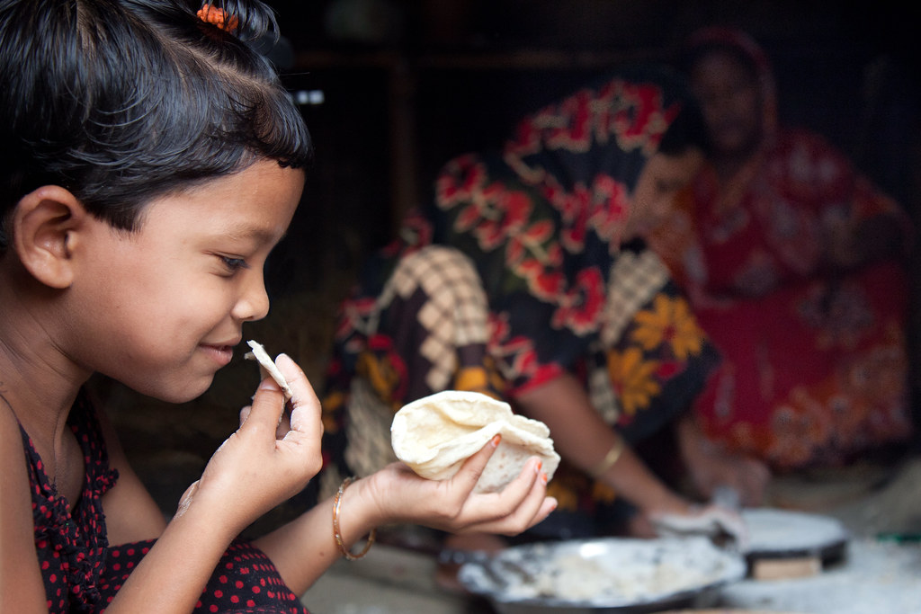 A little girl eats a freshly made roti while the women of her family prepare more, at her home in the village of Chapor, in the district of Dinajpur, Bangladesh. (Photo: S. Mojumder/Drik/CIMMYT)
