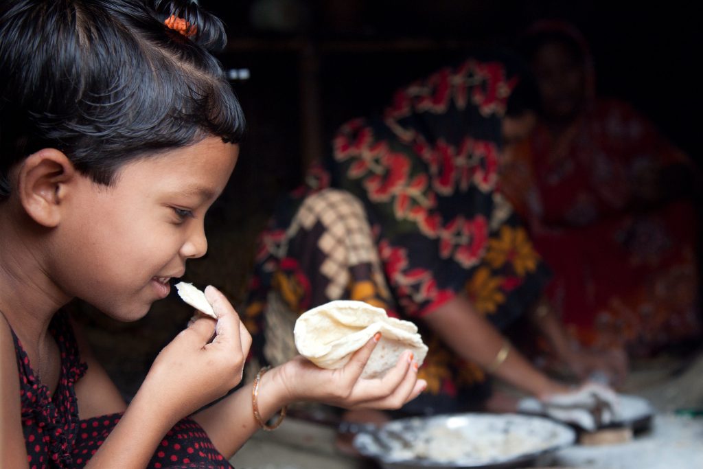 A little girl eats a freshly-made roti while the women of her family prepare more, at her home in the village of Chapor, in the district of Dinajpur, Bangladesh. (Photo: S. Mojumder/Drik/CIMMYT)